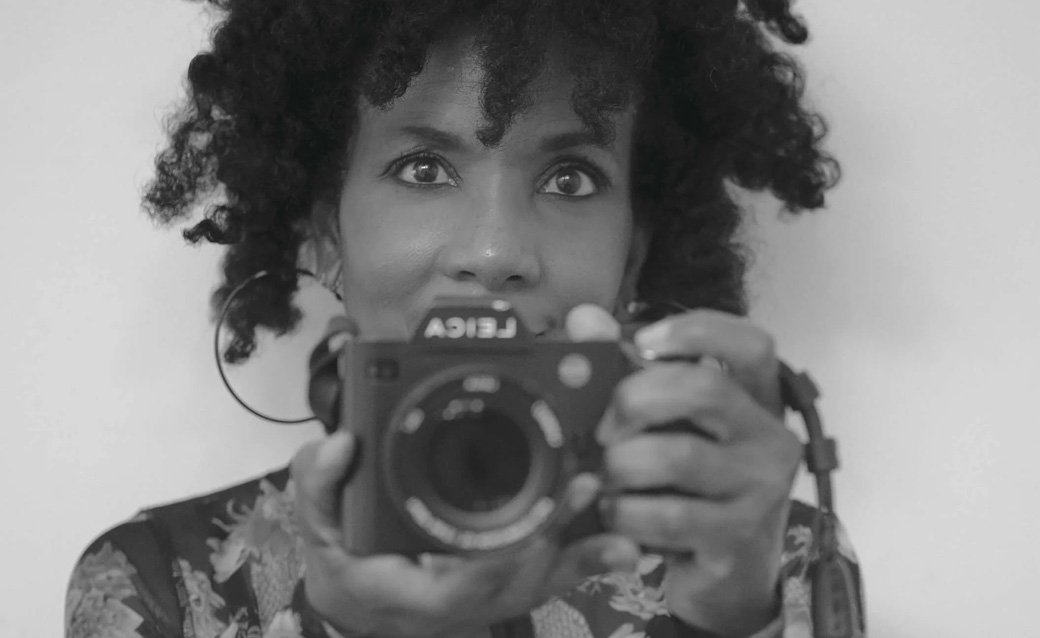 a detail of a black and white image of Sheila Pree Bright in which she holds a camera partially in front of her face, pointing it at the viewer