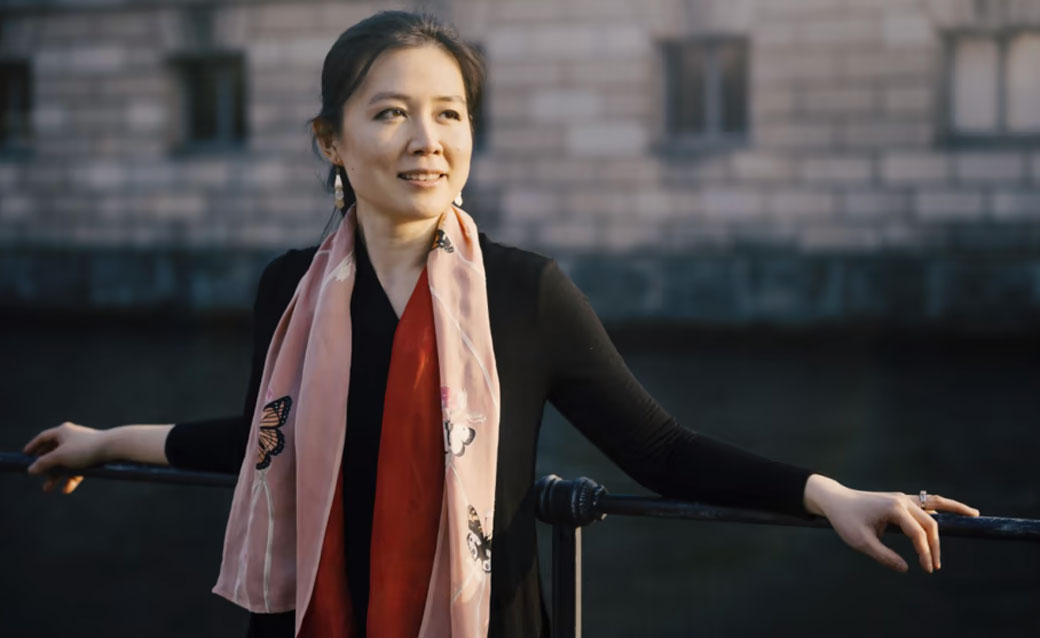 Pianist Amy Yang, leaning against a railing with her arms spread and resting on it. She wears mostly black (with a pink scarf) and has her dark hair pulled back.